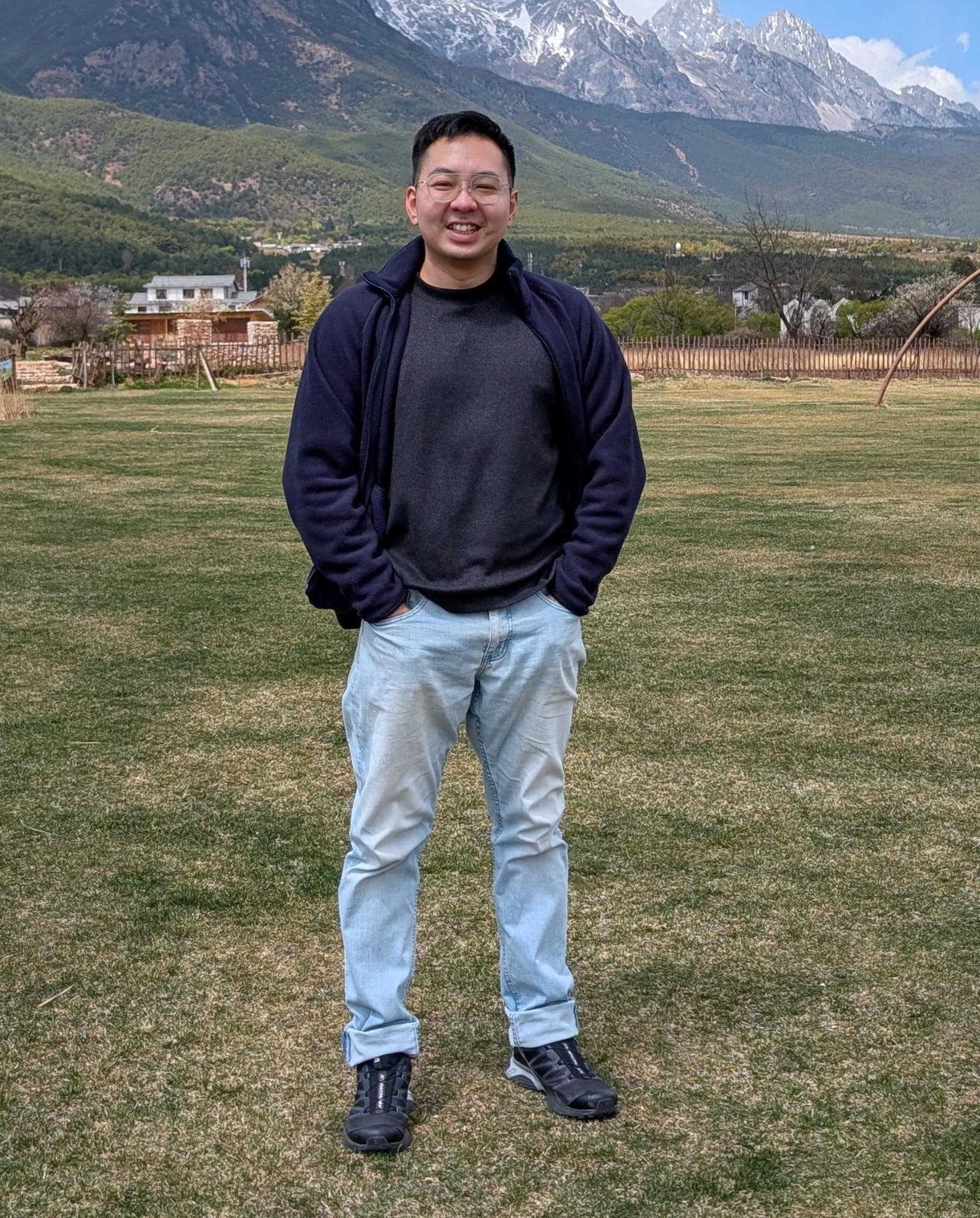 Smiling man in a blue jacket stands in a field before towering snow-capped mountains.