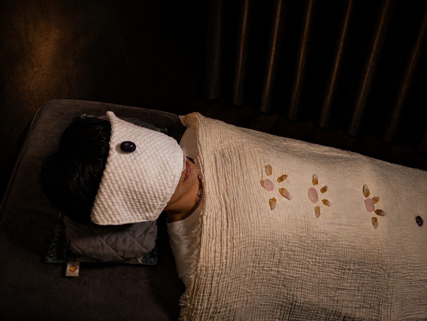 Person wearing an eye mask with crystals arranged on a white blanket for crystal healing.